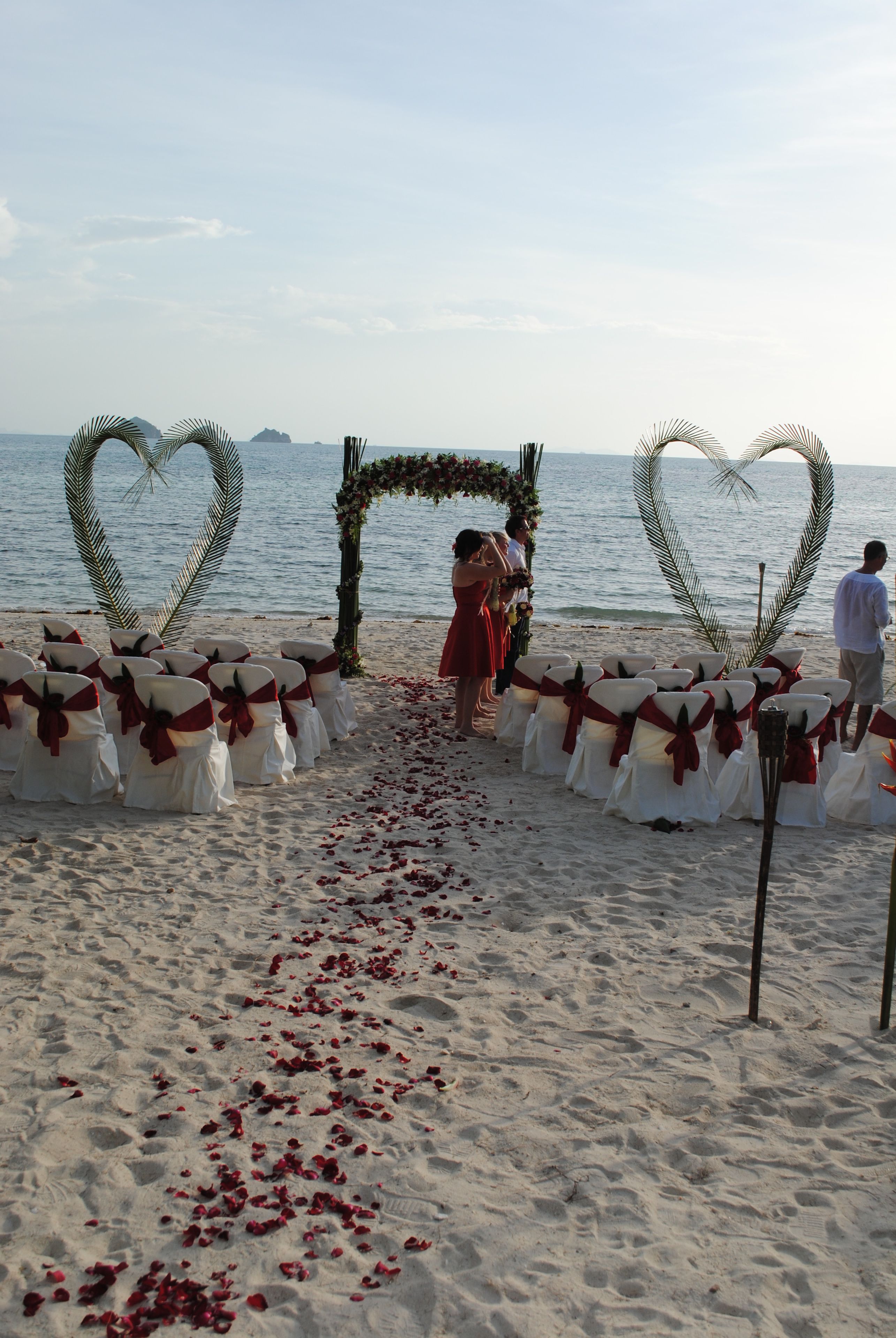 A beach wedding setup with heart-shaped decorations and white chairs with red sashes.