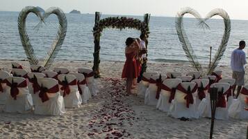 A beach wedding setup with heart-shaped decorations and white chairs with red sashes.