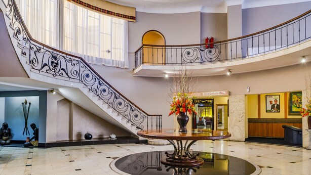 Um hall imponente com uma mesa redonda central, uma escadaria com corrimãos elaborados e um balcão de recepção.