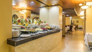 Uma área de buffet com diversos pratos, incluindo frutas e saladas, e um espaço de refeições com mesas e cadeiras.
