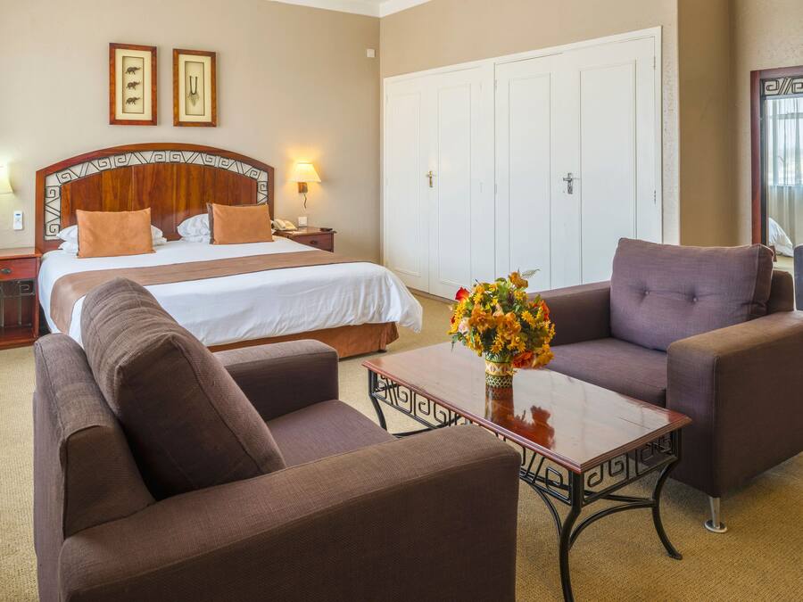A hotel room with a large bed, two brown armchairs, a wooden coffee table with a vase of flowers, a nightstand with a lamp, and a wardrobe.