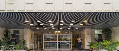 A hotel entrance with a modern design, featuring a glass door, a staircase leading up, and a covered area with a patterned ceiling.