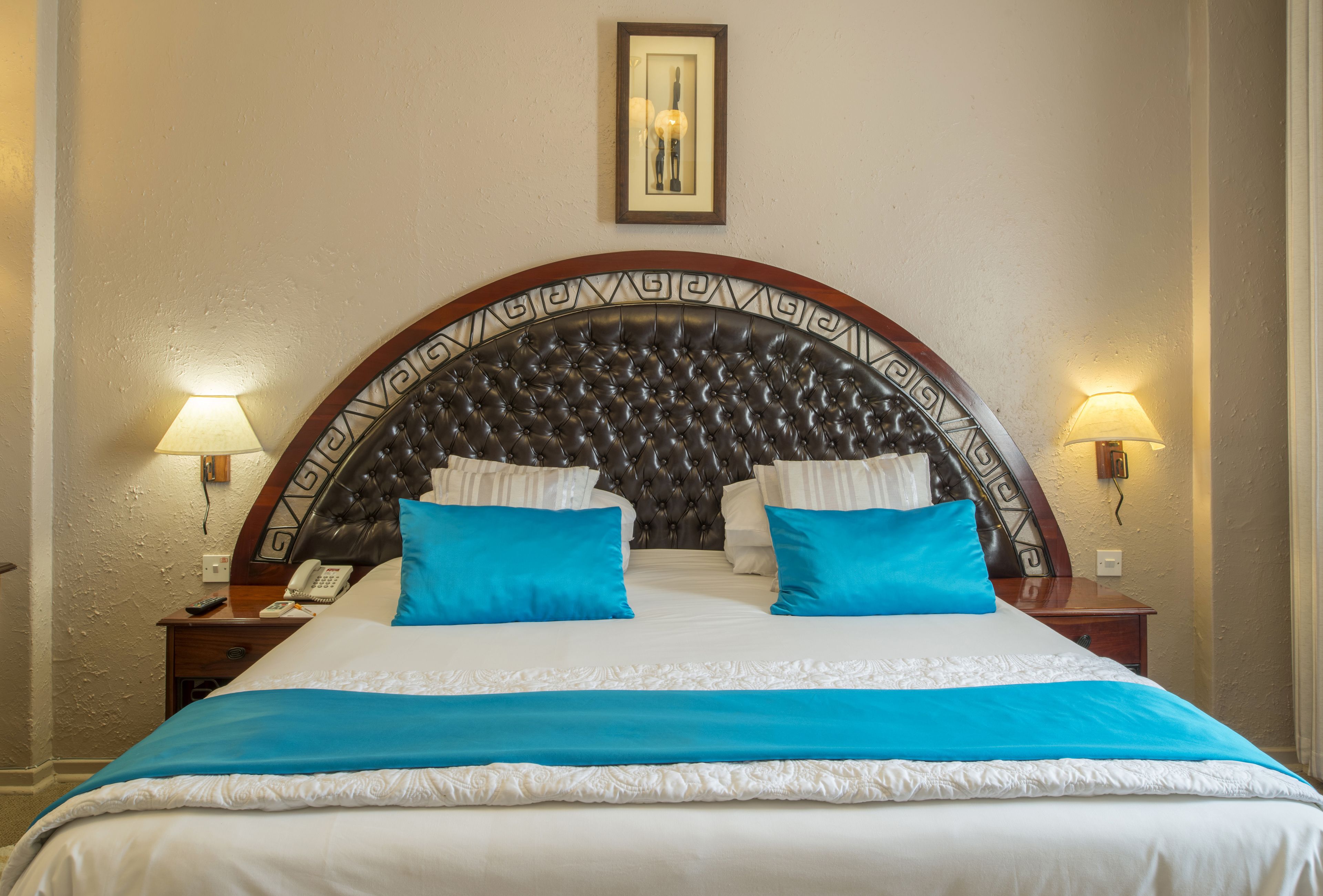 Habitación de hotel con cabecera capitonada, dos lámparas de noche, teléfono y cama con almohada azul decorativa.