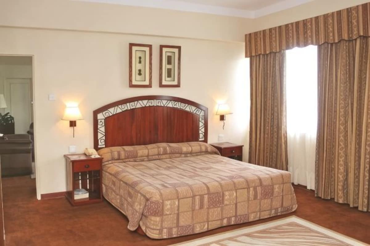 A hotel room with a large bed, two bedside tables, a headboard, two wall-mounted lamps, and a window with curtains.