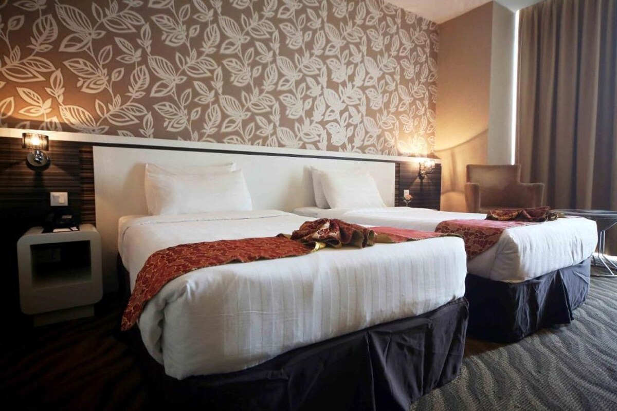 A hotel room with two beds, a patterned wall, bedside lamps, a nightstand, and a chair.