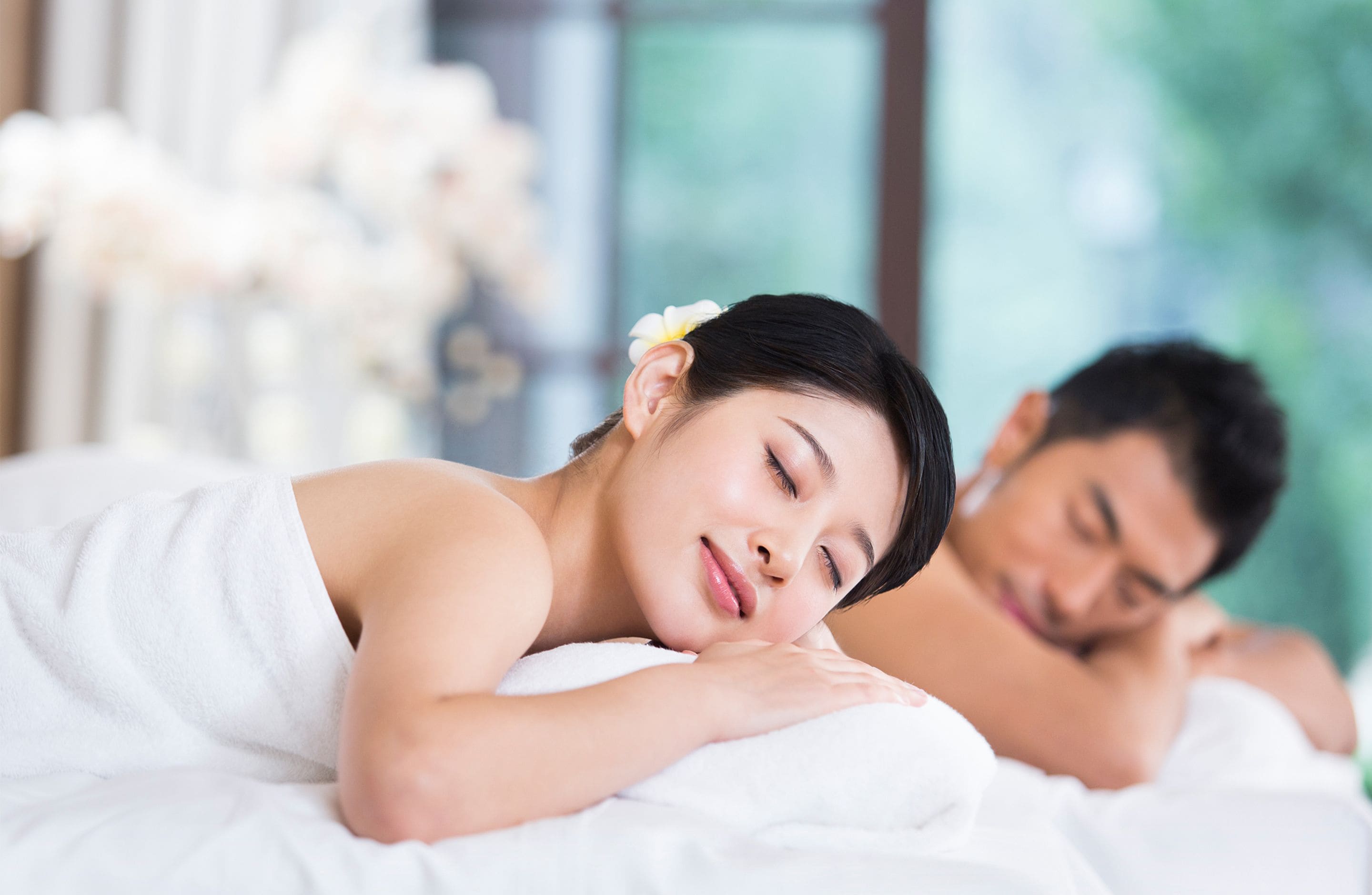 A spa setting with two individuals lying on separate beds, one with a flower in their hair.