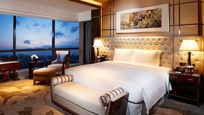 A spacious bedroom with a large bed, two bedside tables with lamps, a chair, and a view of the city at dusk. - Hilton Foshan (Foshan)
