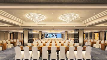 A large conference room with rows of tables and chairs, a stage with a large screen, and chandeliers on the ceiling.
