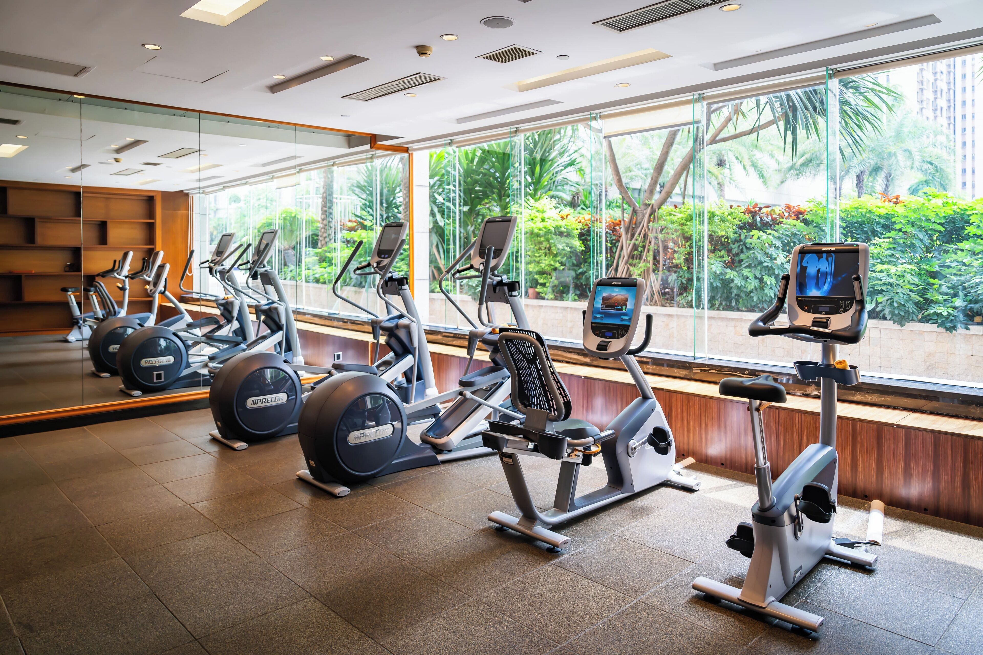 A gym with various exercise machines, large windows with a view of greenery, and a modern interior design.