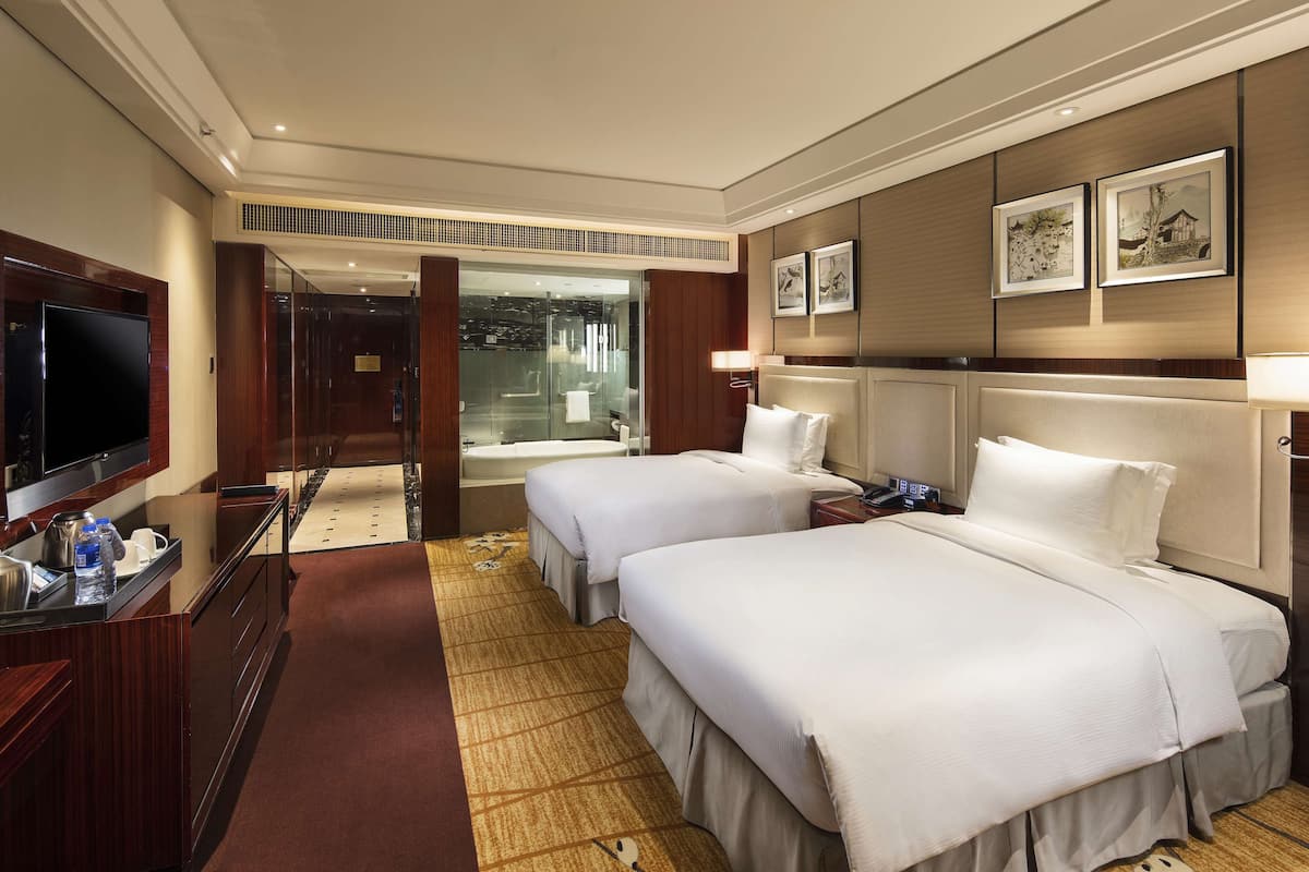 Habitación de hotel con dos camas, televisión, escritorio con computadora y baño visible a través de una puerta de vidrio.