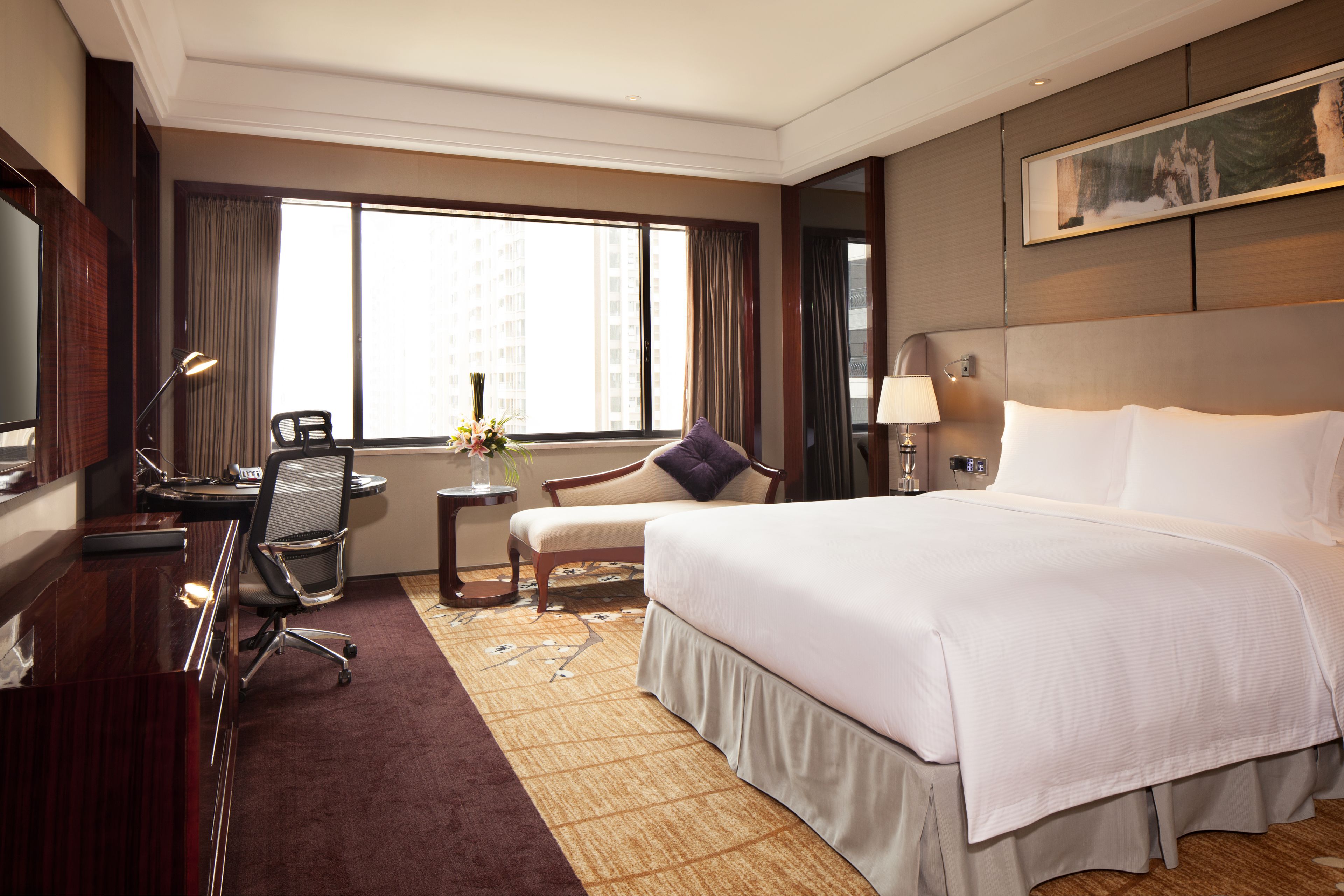 A hotel room with a large bed, a desk with a chair, a small table with a vase, and a large window with curtains.
