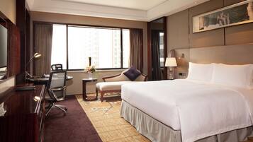 A hotel room with a large bed, a desk with a chair, a small table with a vase, and a large window with curtains.