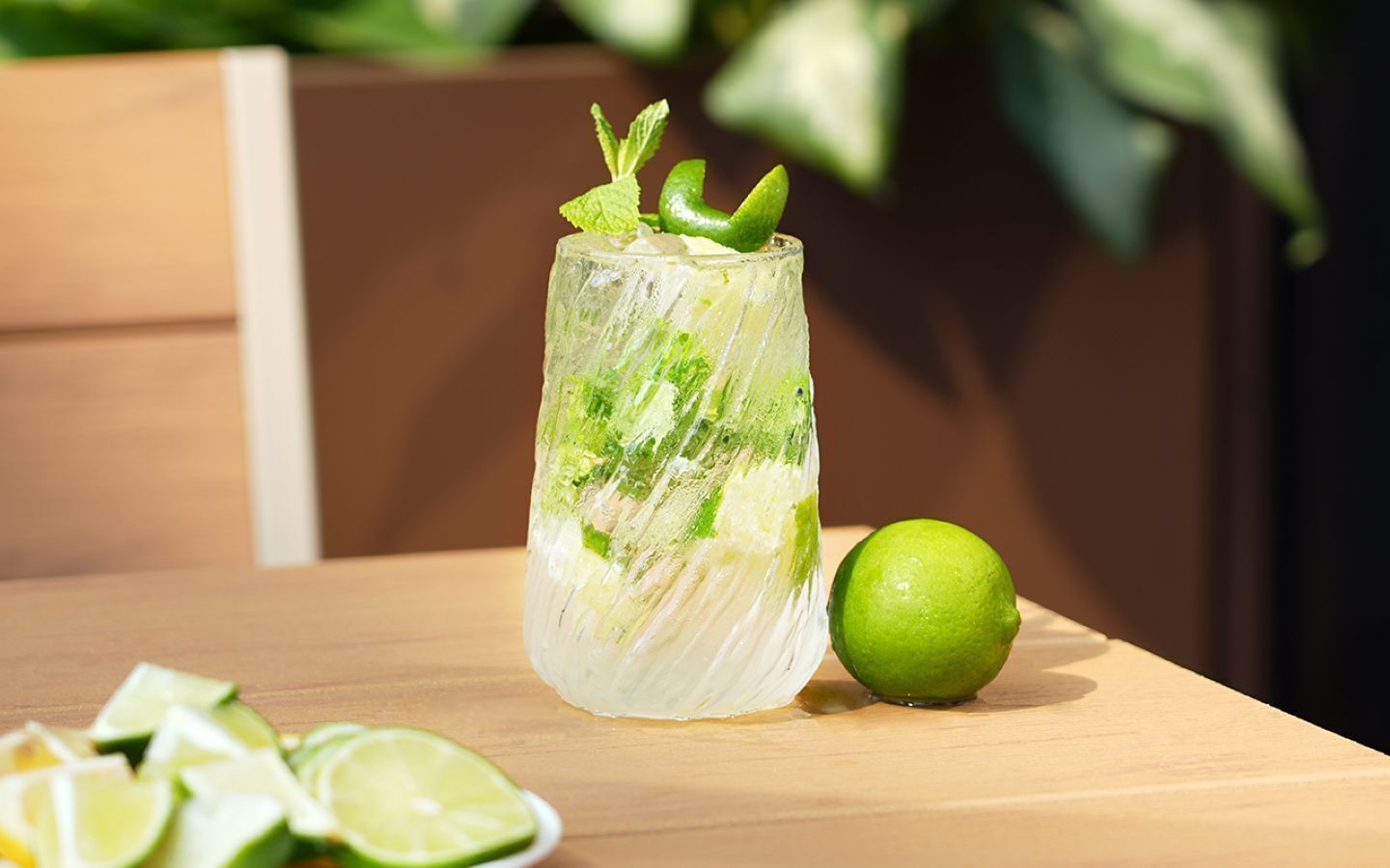 Ett glas med en klar dryck, garnerad med lime och mynta, en hel lime och limeskivor på ett träbord.
