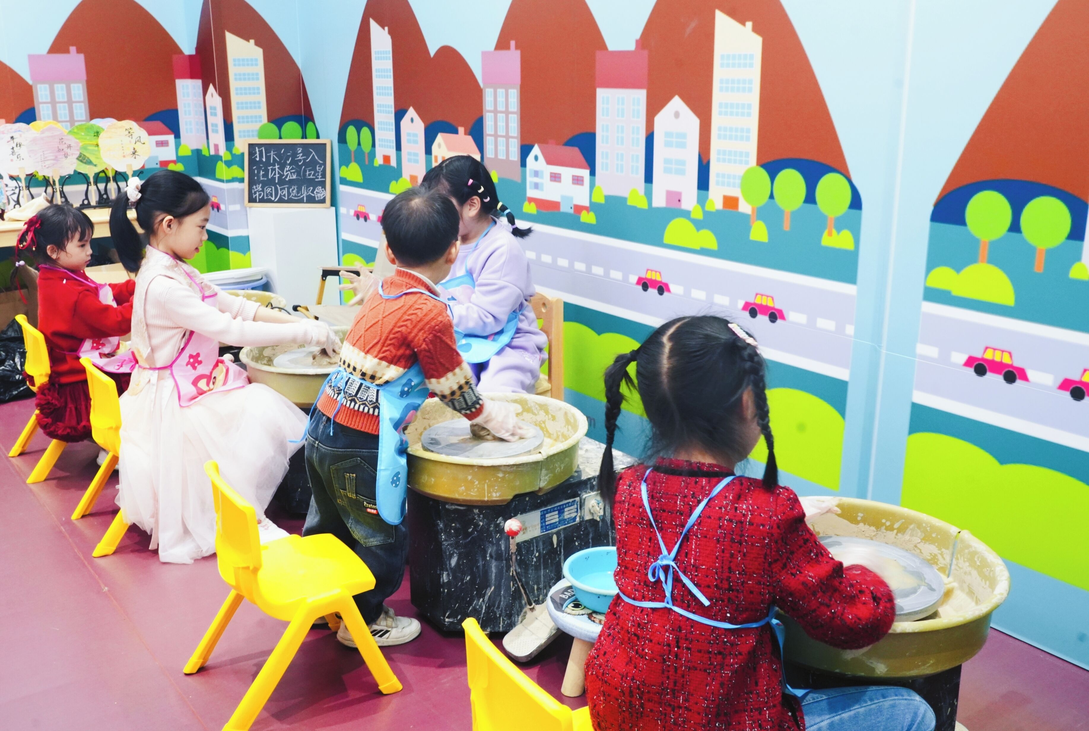 Des enfants jouent avec de grands récipients dans une pièce colorée dont un mur est décoré d’une fresque représentant un paysage urbain.