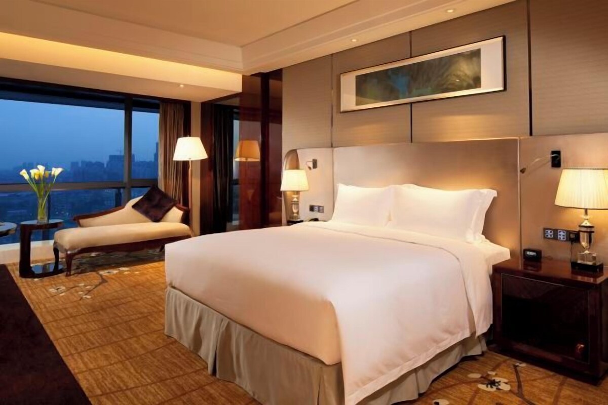 Quarto de hotel com uma cama grande, área de estar com uma poltrona, escrivaninha e vista para a cidade através de amplas janelas.