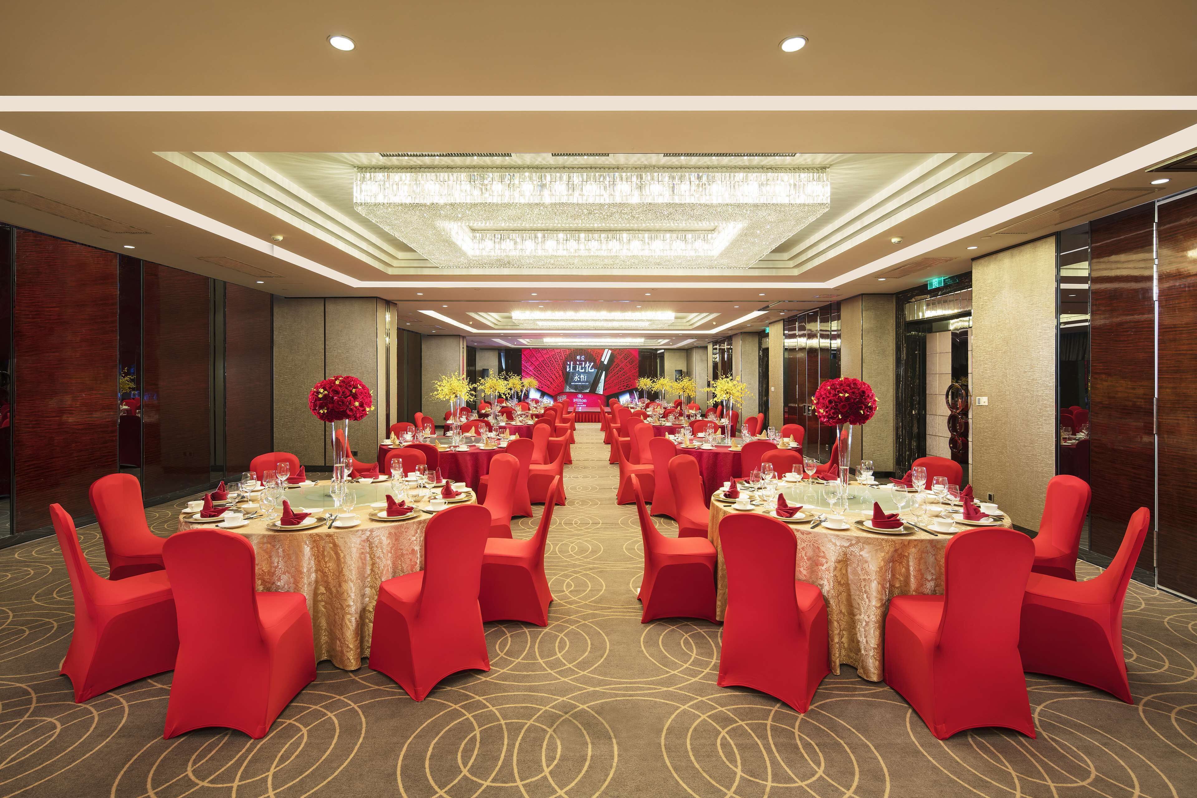 A banquet hall with multiple round tables, red chairs, and chandeliers.