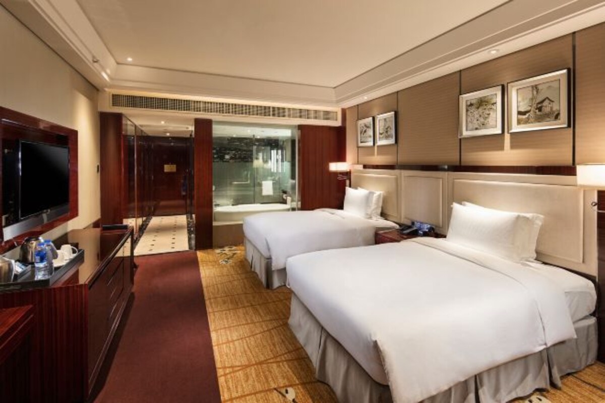 Habitación de hotel con dos camas, televisión, escritorio con computadora y baño visible a través de una puerta de cristal.