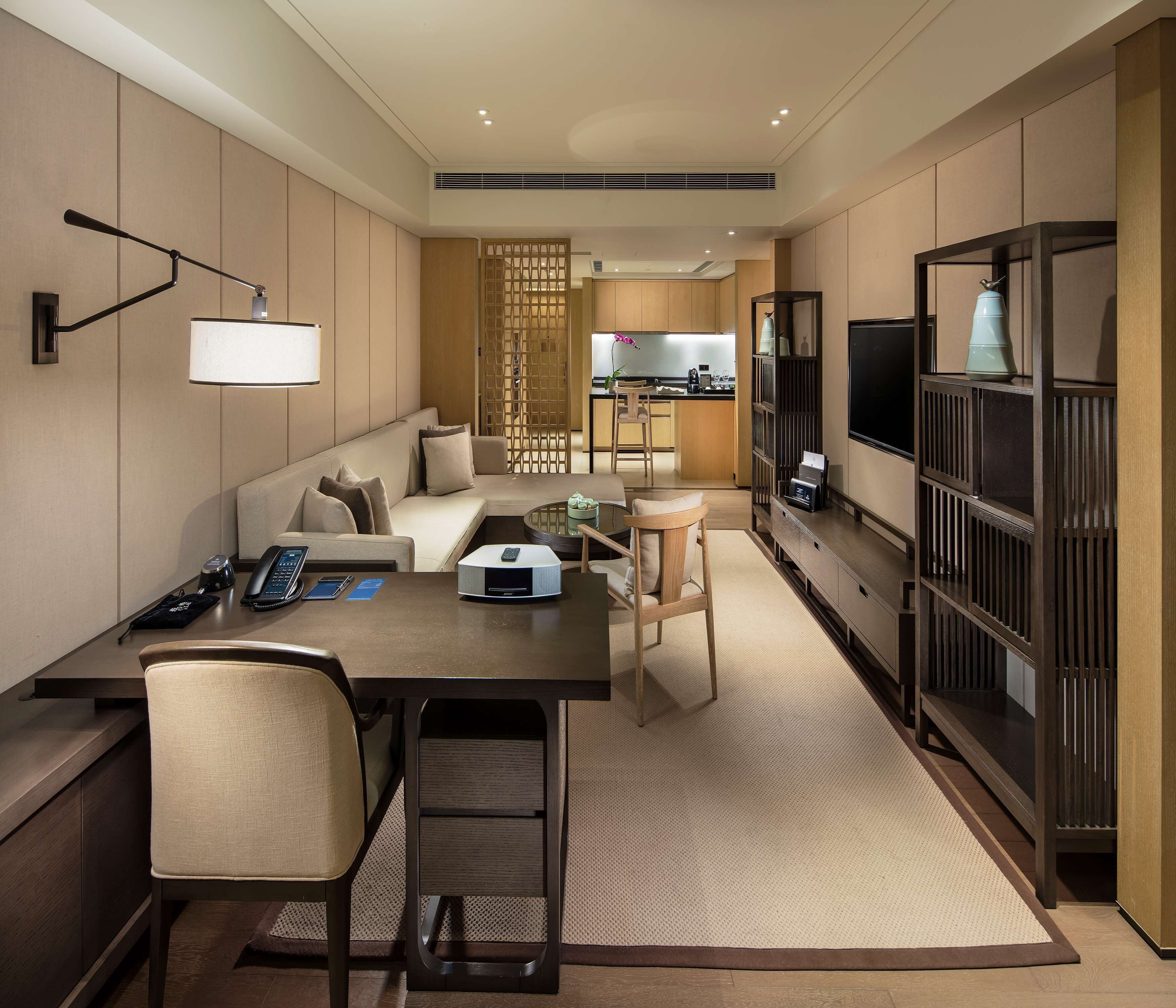 A modern hotel room with a sofa, a dining table, a TV, and a kitchen area in the background.