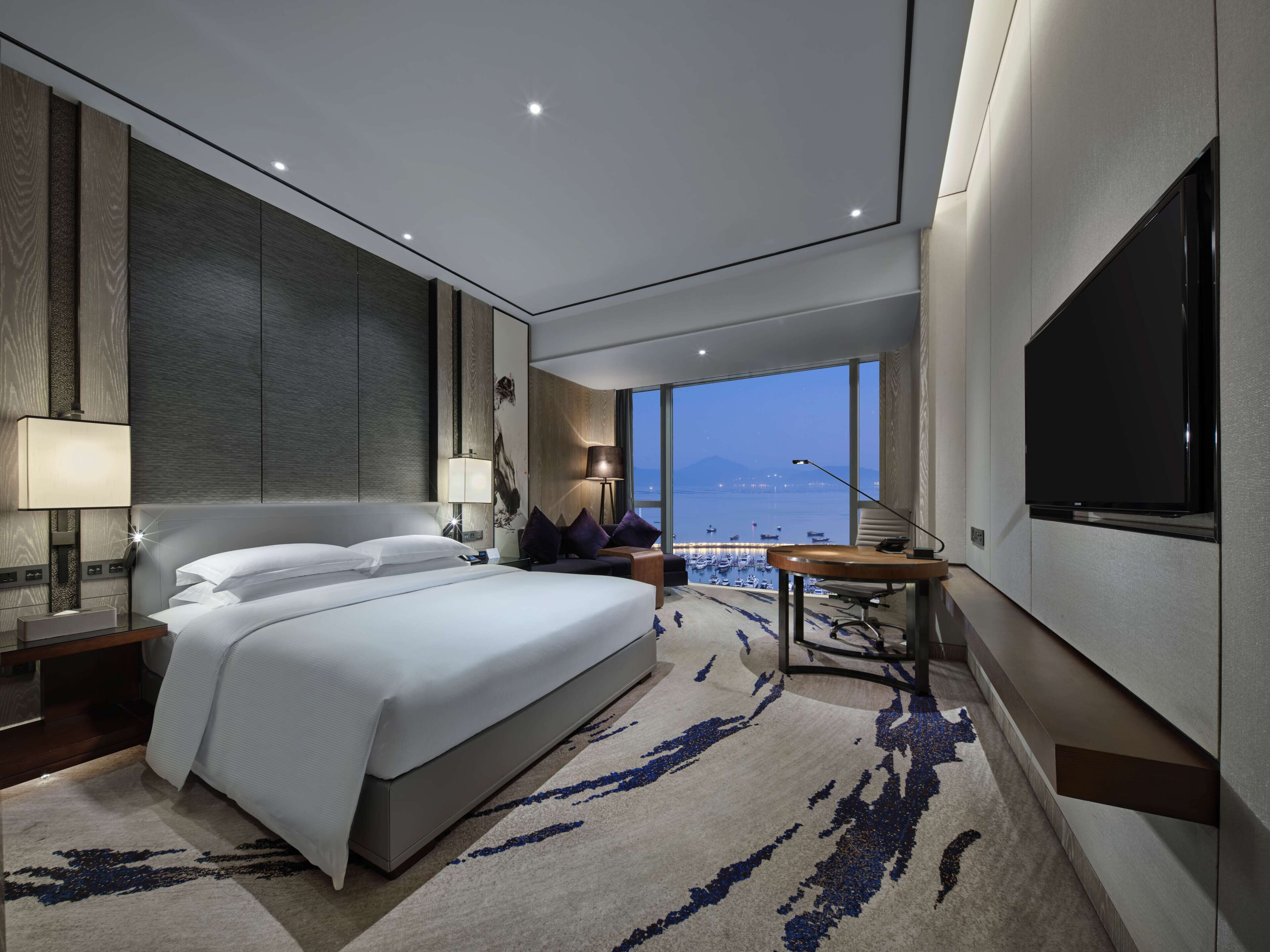 A modern hotel room with a large bed, a desk, a TV, and a view of the sea.