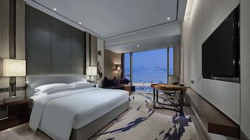 A modern hotel room with a large bed, a desk, a TV, and a view of the sea.