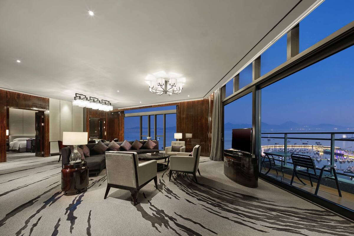 Quarto de hotel espaçoso com uma grande janela que oferece vista para um porto de iates e o horizonte da cidade.