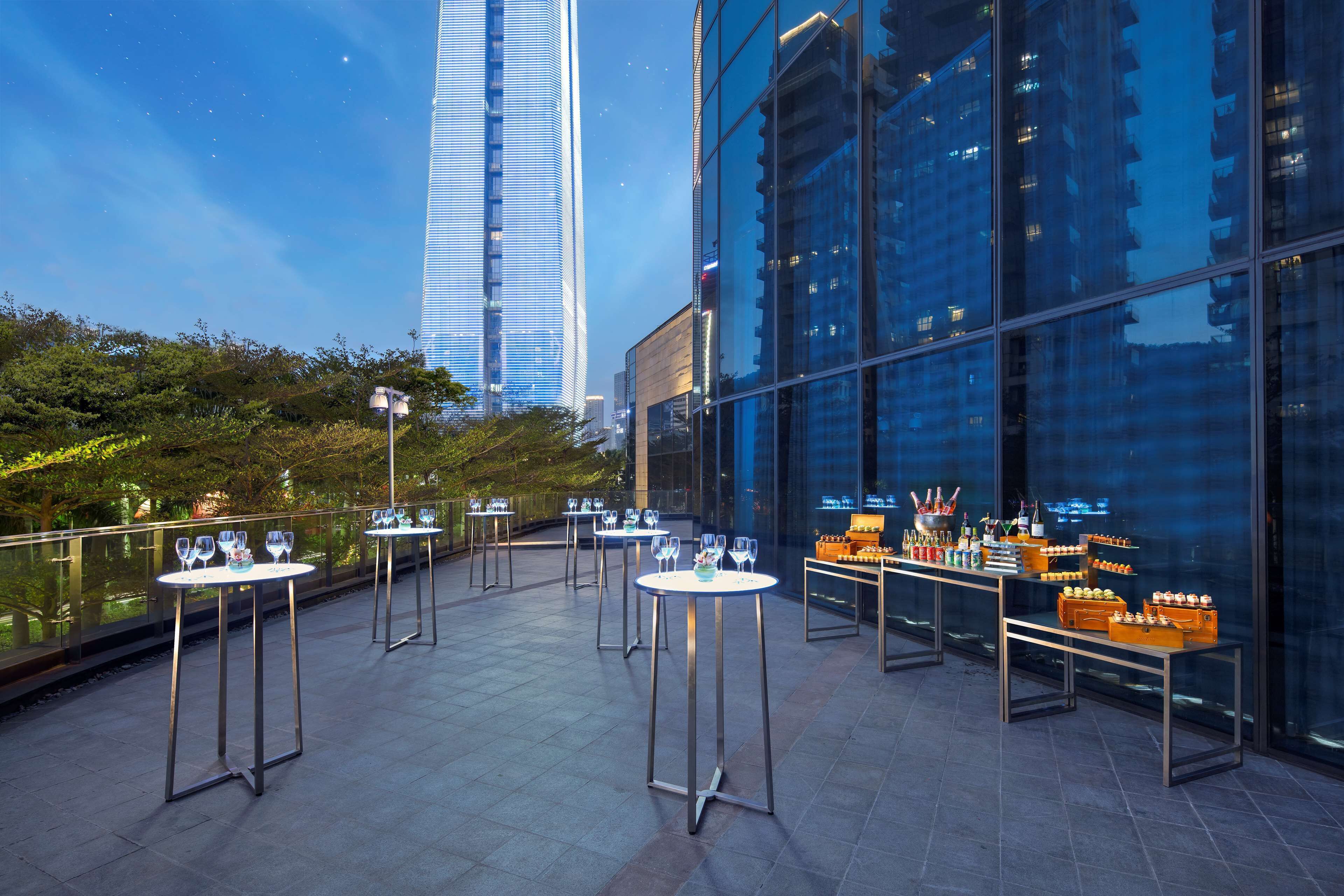 An outdoor terrace with tables and chairs, a glass railing, and a view of tall buildings.