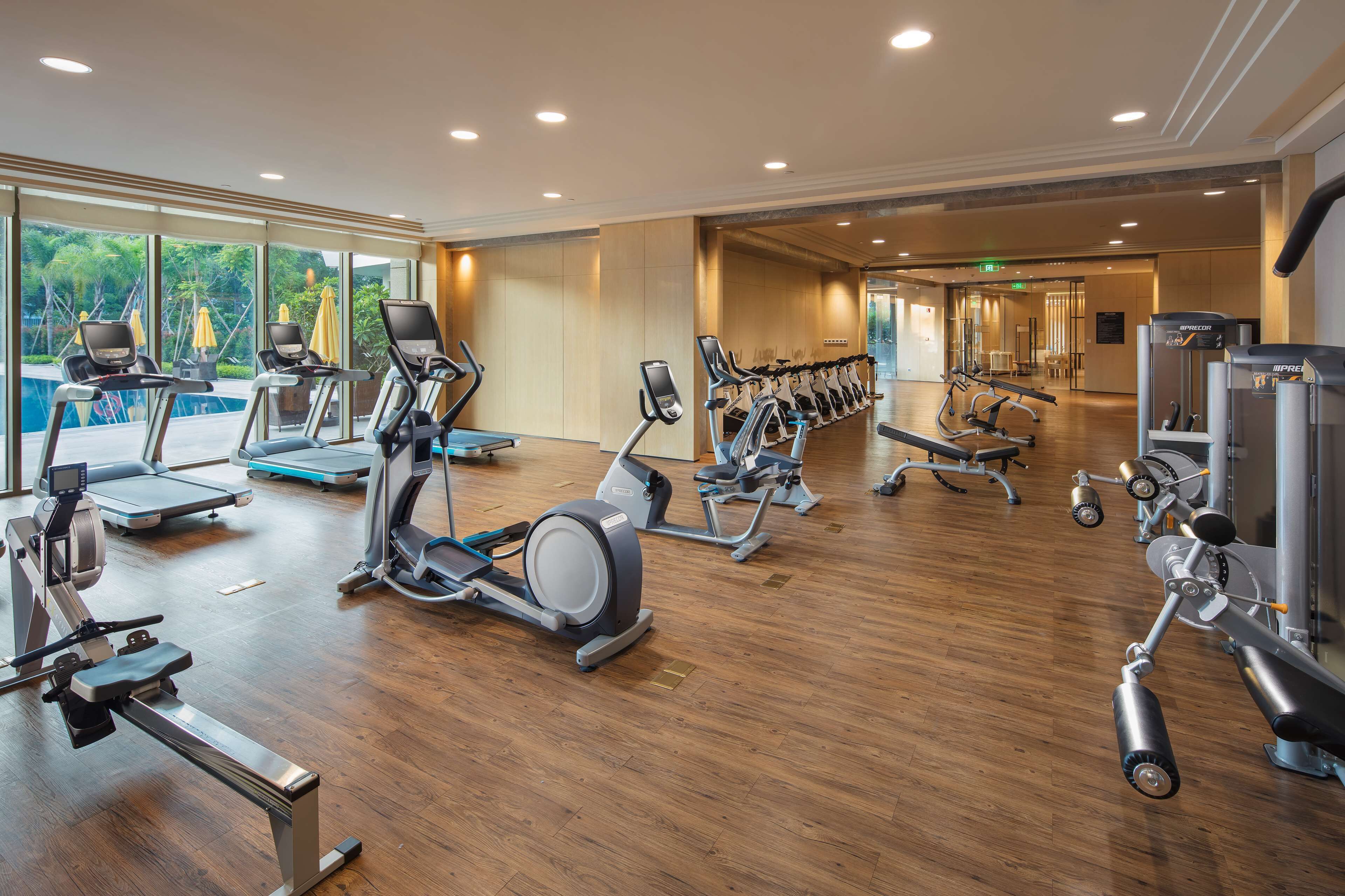 A gym with various cardio and strength training equipment, large windows, and a wooden floor.
