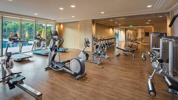 A gym with various cardio and strength training equipment, large windows, and a wooden floor.