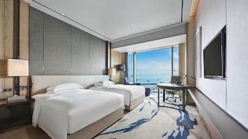 A hotel room with two beds, a desk, a chair, a TV, and a large window with a view.