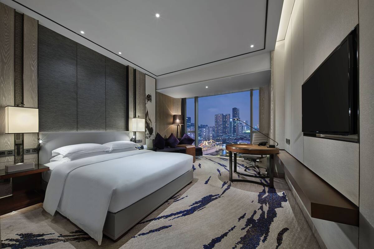 Quarto de hotel moderno com uma cama grande, uma escrivaninha, uma televisão e vista para a cidade.