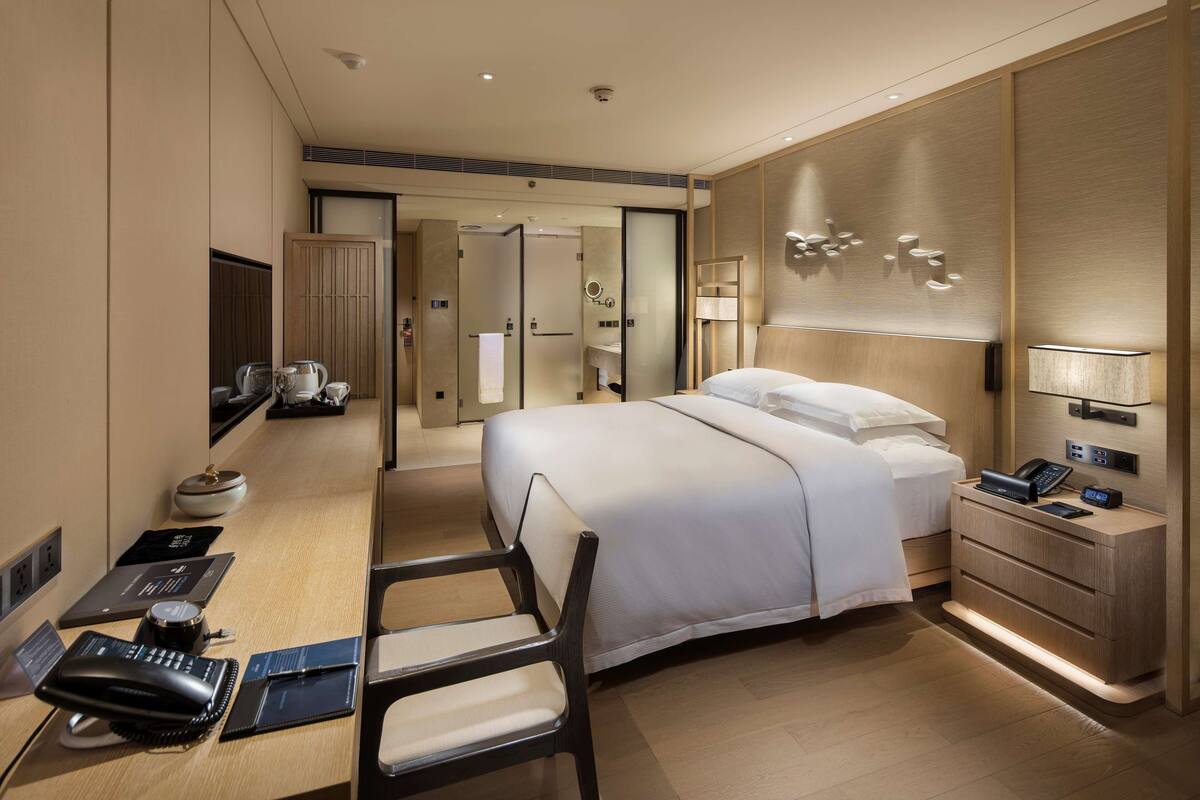 Quarto de hotel moderno com uma cama grande, uma escrivaninha com um laptop, uma cadeira, uma mesa de cabeceira com uma luminária e um guarda-roupa.