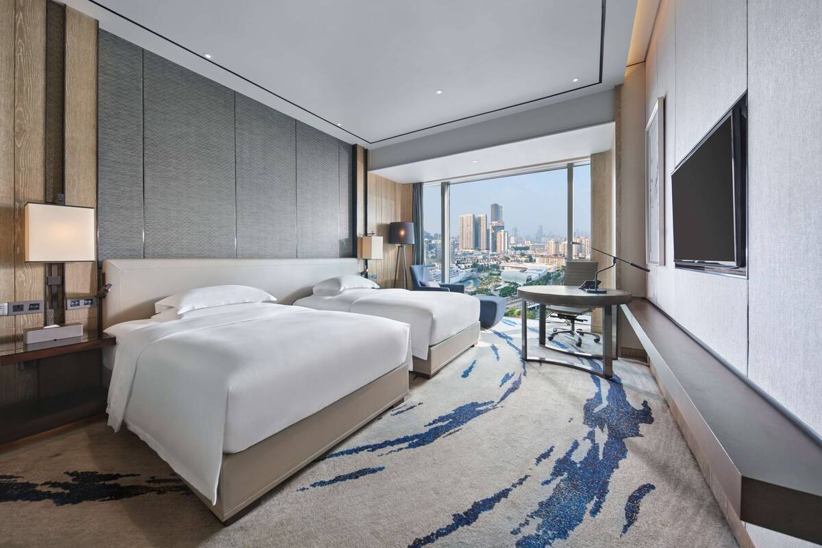 Quarto de hotel moderno com duas camas, uma escrivaninha, uma cadeira, uma janela grande com vista para a cidade e uma TV de tela plana.