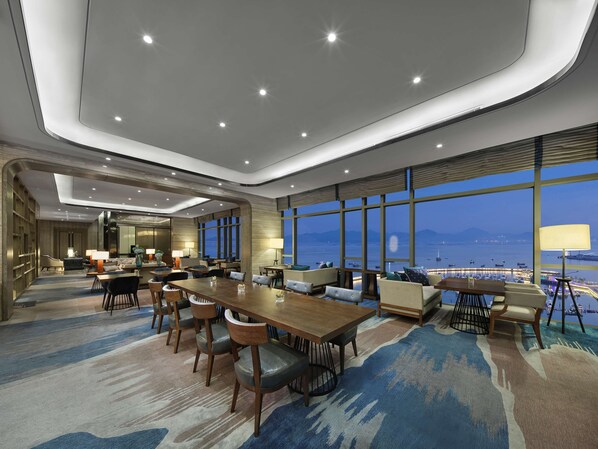 Un hall de hotel espacioso con una gran mesa de comedor, asientos cómodos y vistas al agua y embarcaciones.