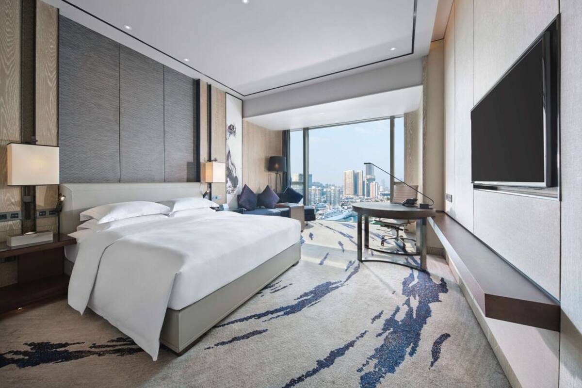 Quarto de hotel moderno com uma cama grande, uma escrivaninha, uma cadeira e vista para a cidade.