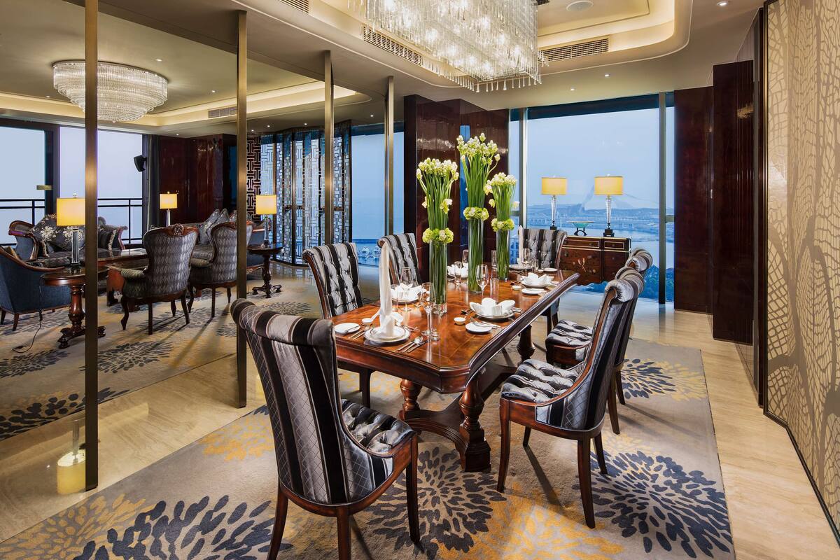 Uma sala de jantar luxuosa com um candelabro grande, cadeiras elegantes e vista para a paisagem urbana através de grandes janelas.