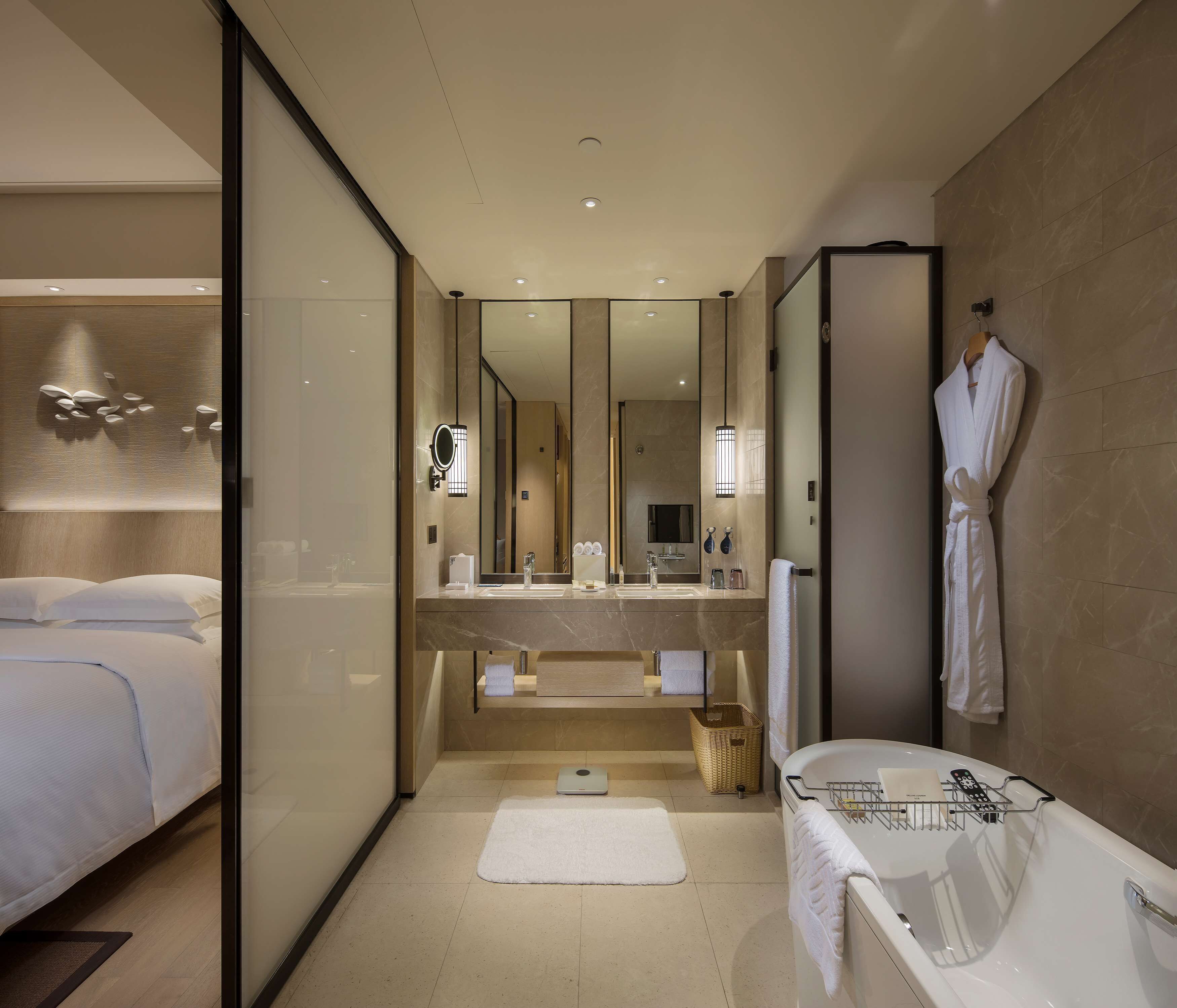 A modern hotel bathroom with a large mirror, a bathtub, a vanity with a sink, and a robe hanging on the wall.