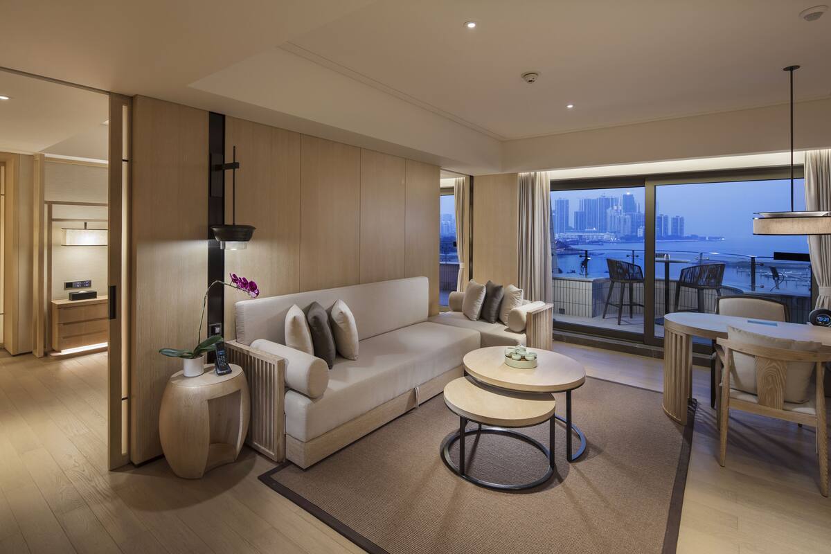 Quarto de hotel moderno com sofá, duas mesas redondas, vista para a cidade e varanda com mesa e cadeiras.