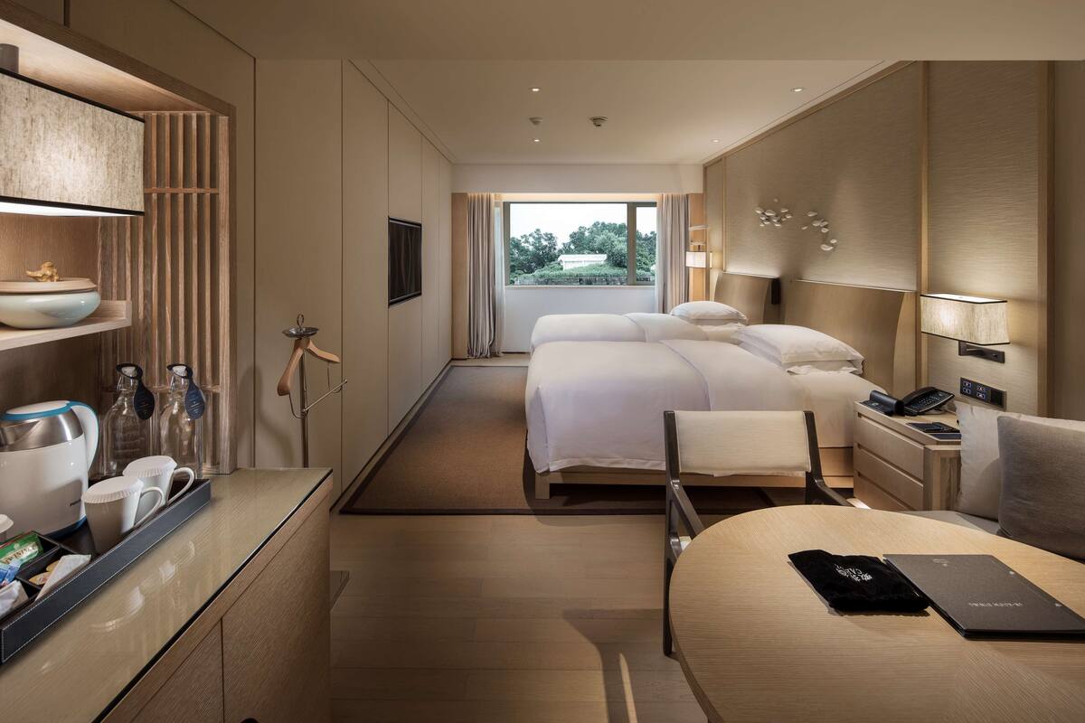 Quarto de hotel moderno com uma cama grande, uma escrivaninha com um laptop, um espaço para café e vista para o verde lá fora.