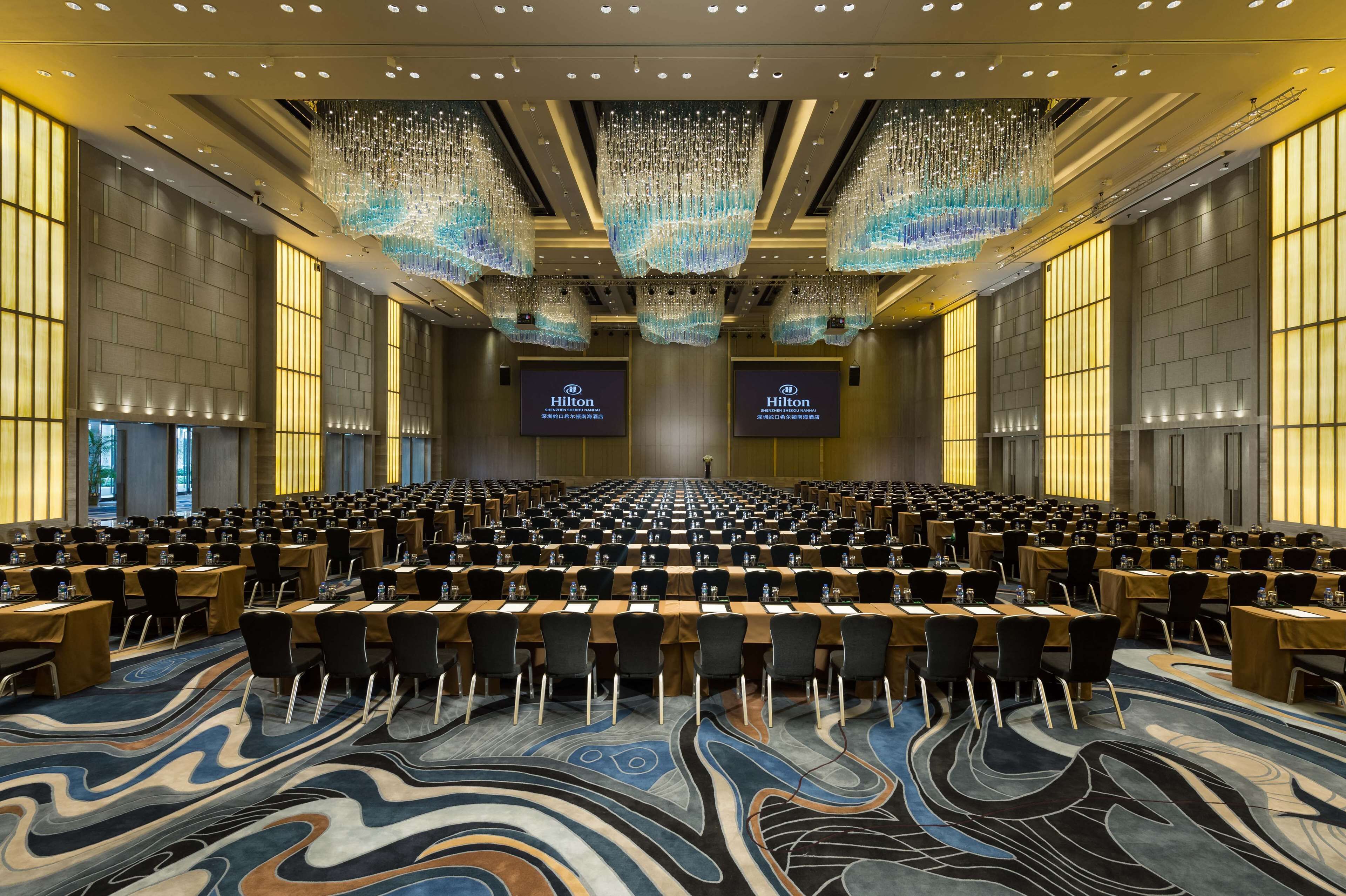 Un amplio salón de conferencias con filas de mesas y sillas, candelabros y pantallas que muestran el logotipo de Hilton.
