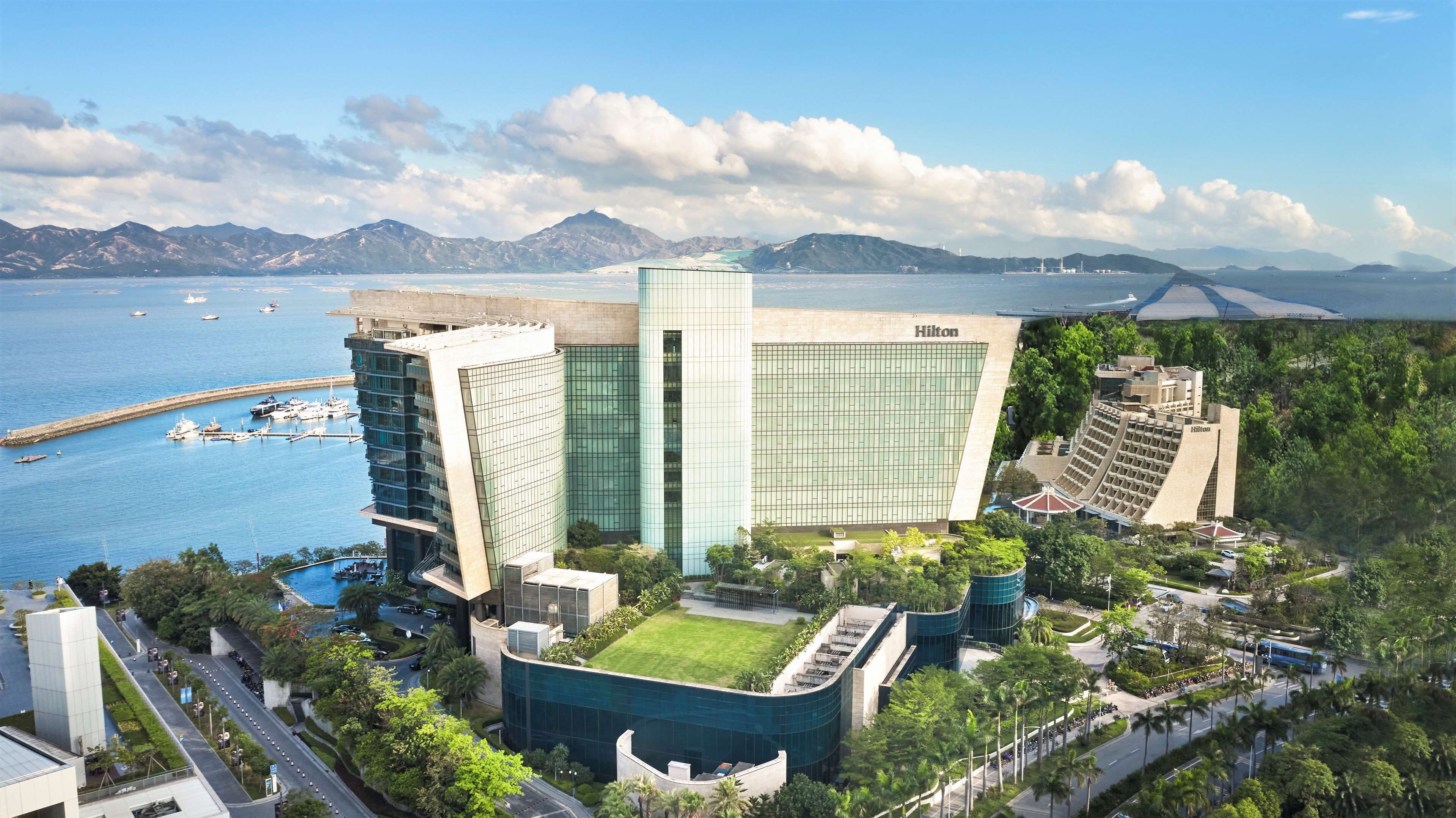 Un hôtel moderne à la façade vitrée, situé près d’une étendue d’eau, avec des bateaux et des montagnes en arrière-plan.