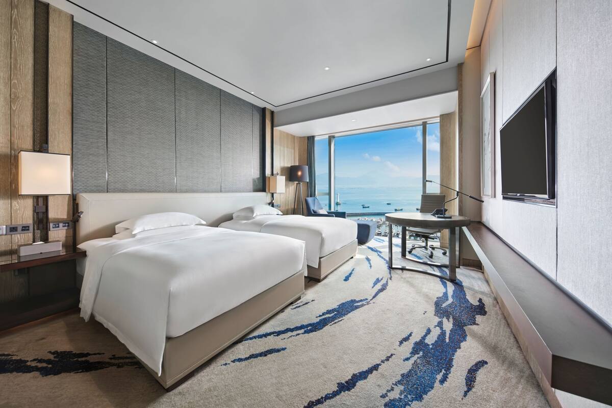 Quarto de hotel moderno com duas camas, uma escrivaninha, uma janela ampla com vista para o mar e uma TV de tela plana.