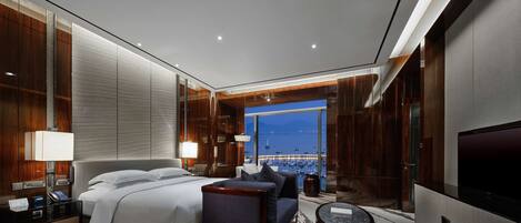 A modern hotel room with a large bed, a sofa, a small round table, and a view of a harbor at night.