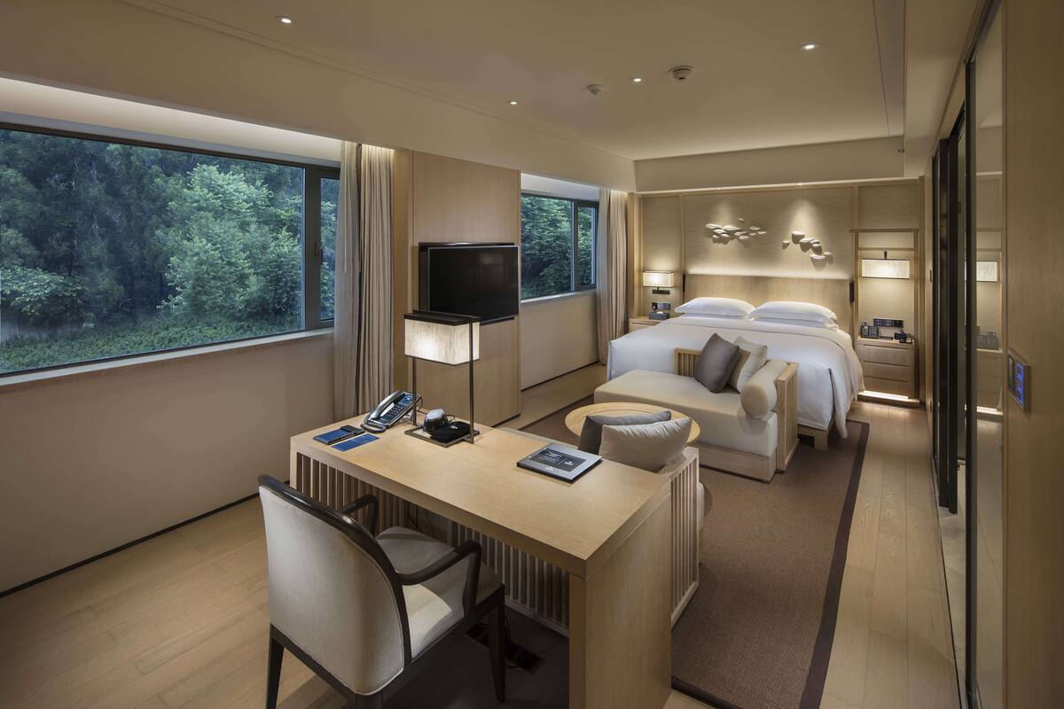 Quarto de hotel moderno com uma janela grande, uma mesa com cadeira, uma cama e uma TV.