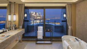 Une salle de bain moderne avec un grand miroir, deux vasques, une baignoire et une vue sur le paysage urbain au crépuscule.