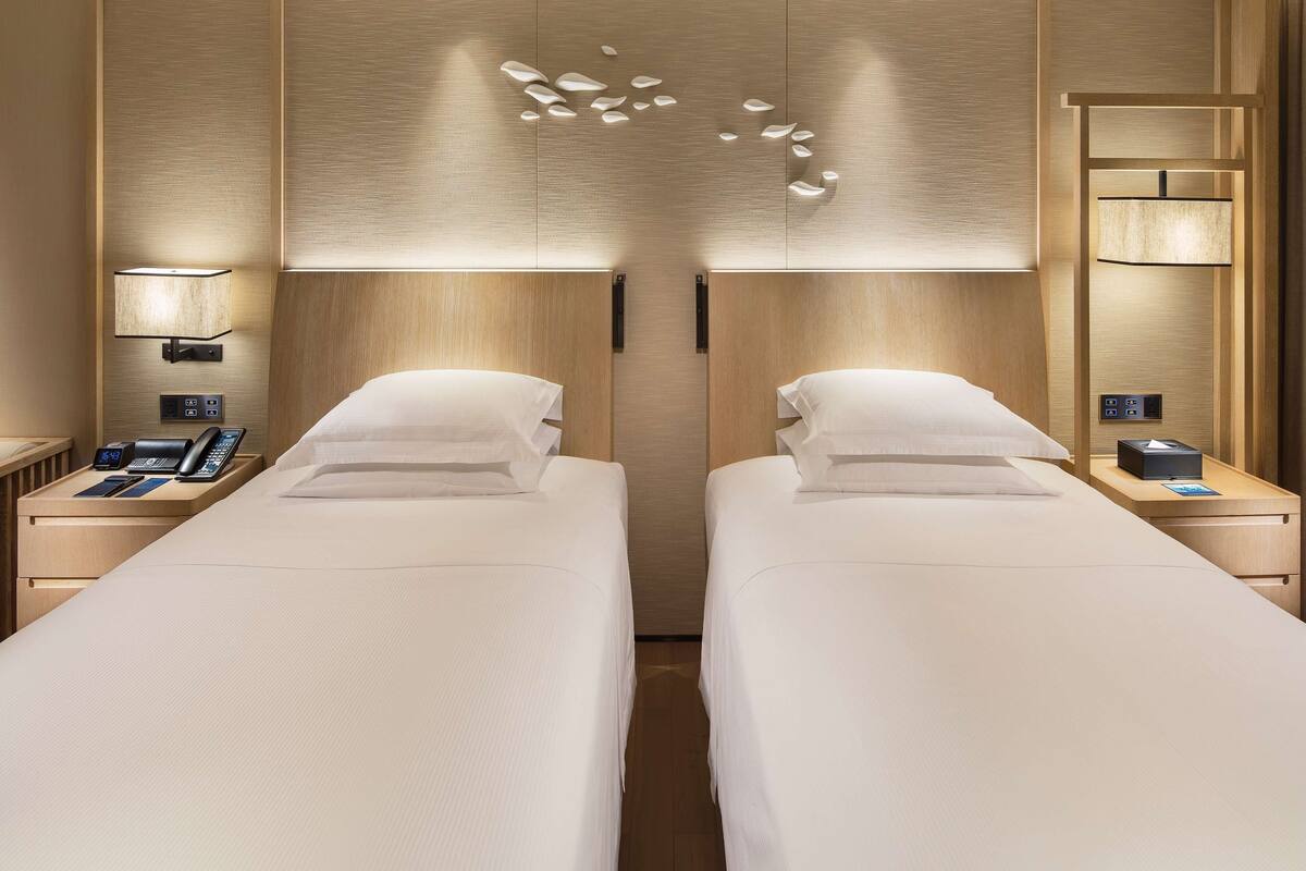 Quarto de hotel com duas camas, mesa de cabeceira com telefone e luminária, e arandela na parede.
