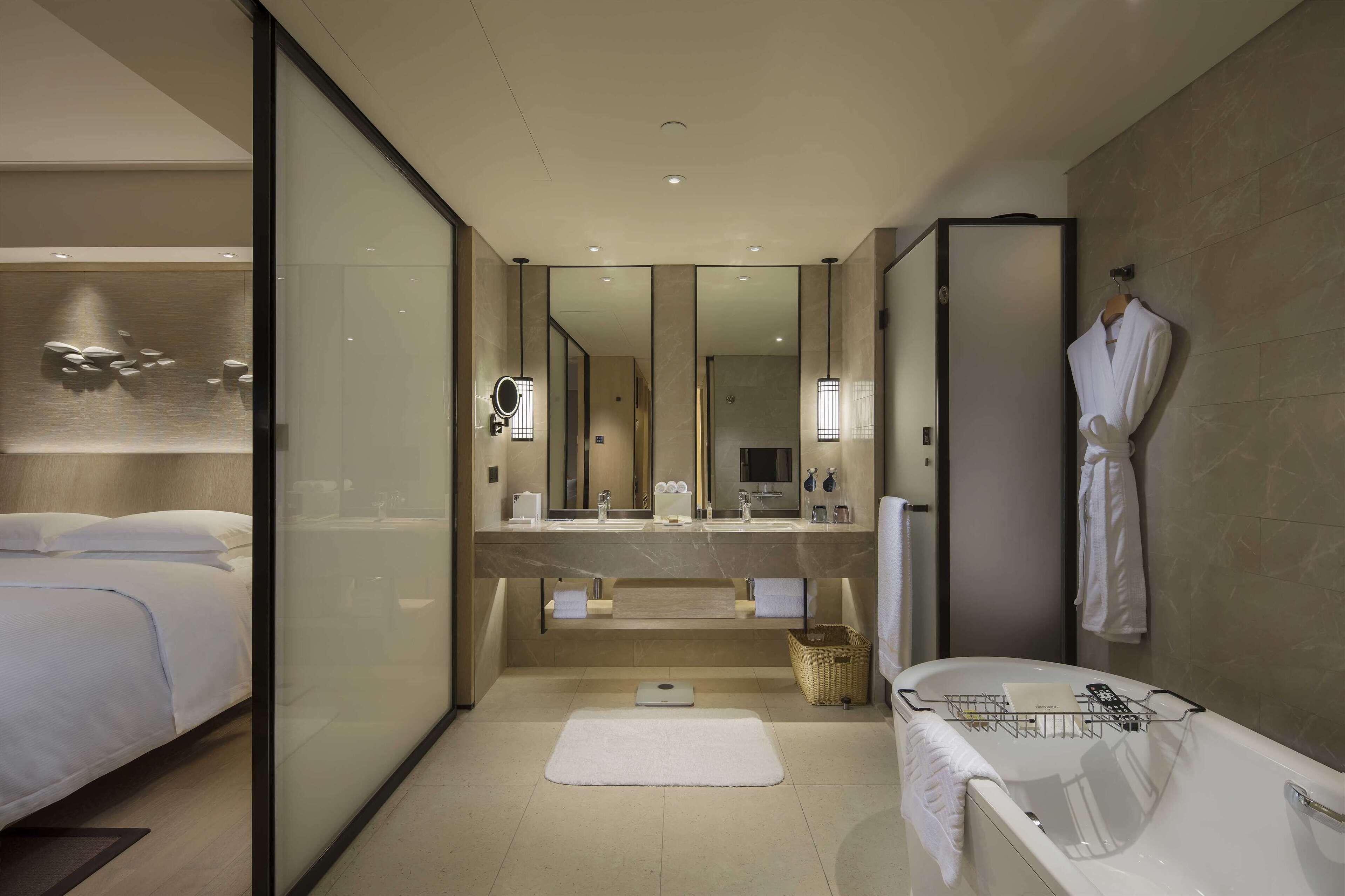 A modern hotel bathroom with a large mirror, a bathtub, a shower, and a glass-enclosed walk-in closet.