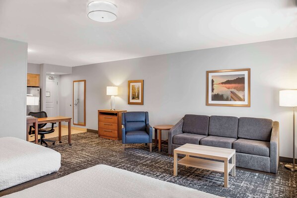A hotel room with a bed, a sofa, a desk with a chair, a small table, a lamp, and a framed picture on the wall.
