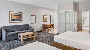 A hotel room with a gray sofa, a wooden coffee table, a bed, a desk with a chair, and a bathroom with a shower and bathtub. - Homewood Suites by Hilton Winnipeg Airport-Polo Park, MB (Winnipeg)