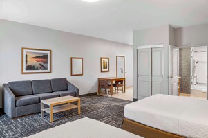 Una habitación de hotel con un sofá gris, una mesa de centro de madera, una cama, un escritorio con una silla y un baño con ducha y bañera.