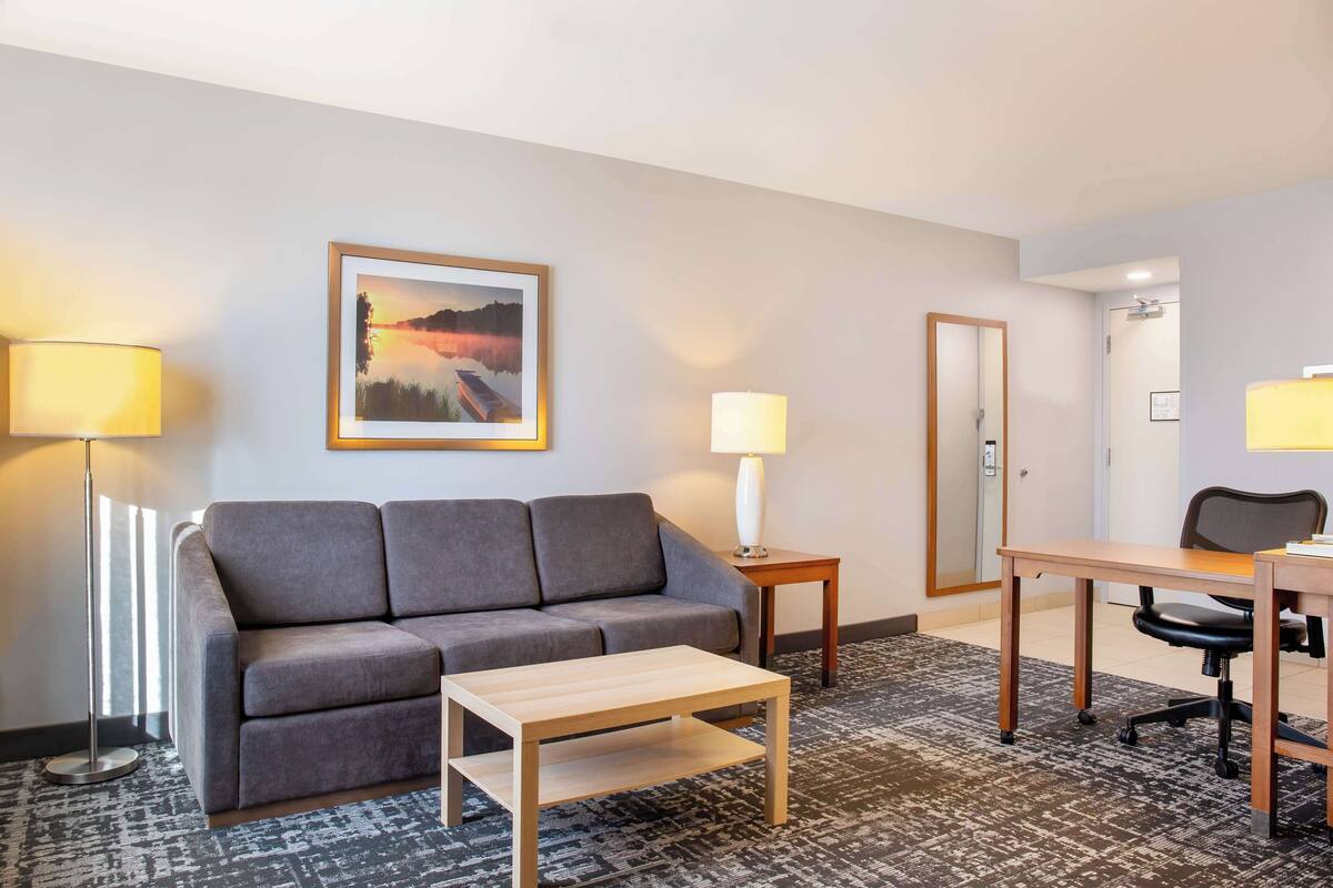 Ein Hotelzimmer mit grauem Sofa, einem kleinen Holztisch, einem Schreibtisch mit Stuhl, zwei Lampen und einem gerahmten Bild an der Wand.